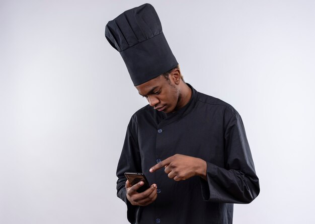 Young confused afro-american cook in chef uniform looks at and points at phone isolated on white wall 