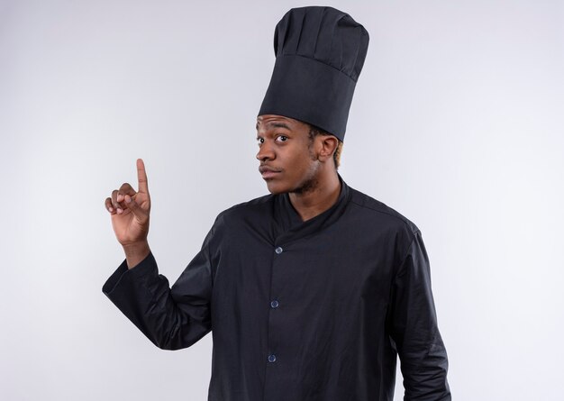 Молодой растерянный афро-американский повар в униформе шеф-повара указывает вверх, изолированные на белой стене
