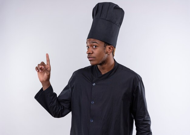 Young confused afro-american cook in chef uniform points up isolated on white wall 