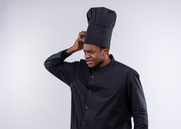 Молодой растерянный афро-американский повар в униформе шеф-повара кладет руку на голову, изолированную на белой стене