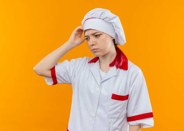 요리사 유니폼에 젊은 혼란 금발 여성 요리사 머리를 보유하고 오렌지 벽에 고립 된 모습