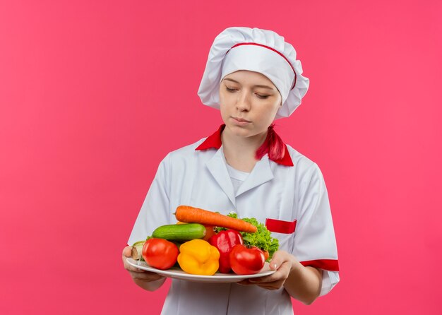 Young confused blonde female chef in chef uniform holds and looks at vegetables on plate isolated on pink wall