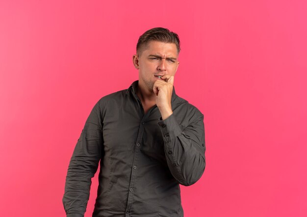 Young confused blonde handsome man puts hand on chin looking at side isolated on pink background with copy space