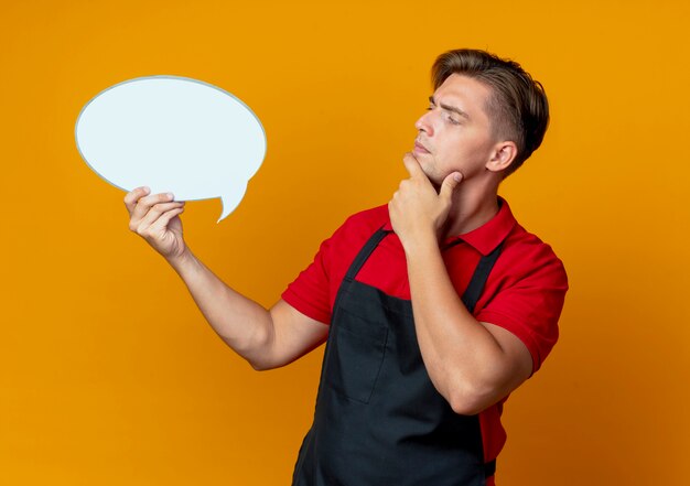 Young confused blonde male barber in uniform holds and looks at chat bubble isolated on orange space with copy space