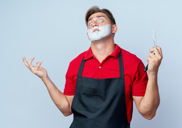 Young confused blonde male barber in uniform smeared face with shaving foam holding straight razor isolated on white space with copy space