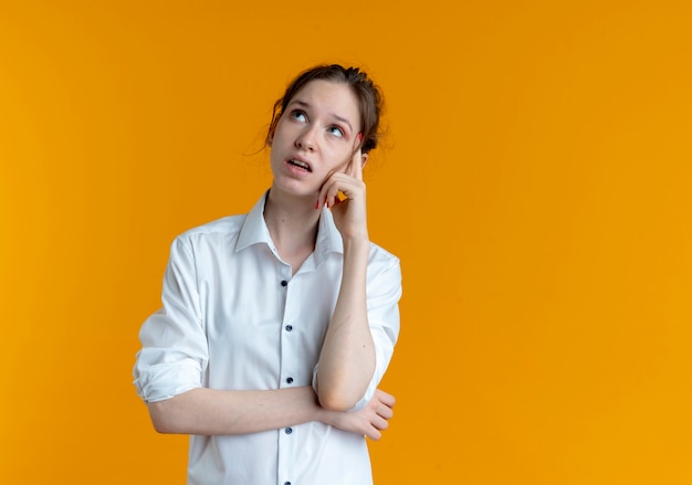 Young confused blonde russian girl puts hand on chin looking up isolated on orange space with copy space