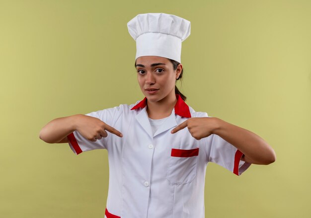 Young confused caucasian cook girl in chef uniform points at herself isolated on green wall with copy space