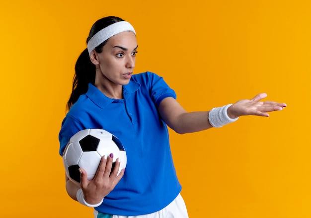 Young confused caucasian sporty woman wearing headband and wristbands holds ball pointing at side isolated on orange background with copy space