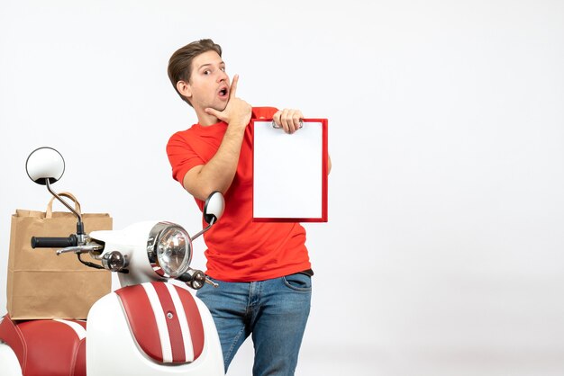 Молодой сбитый с толку курьер в красной форме стоит возле скутера, показывая документ на белой стене