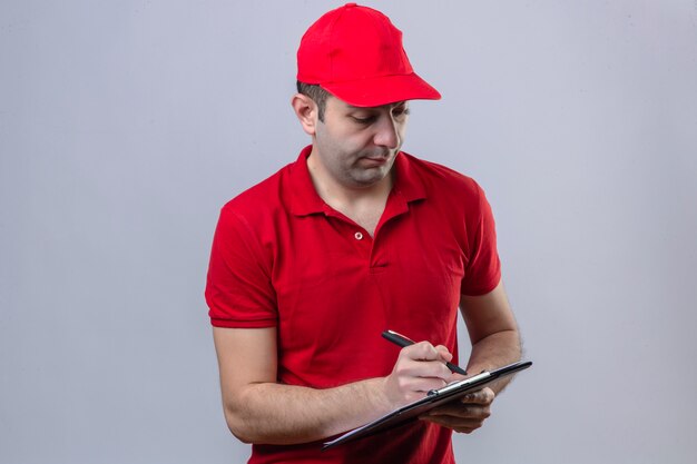 若い混乱赤いポロシャツとキャップクリップボードを保持していると孤立した白い壁に深刻な顔をして空白に何かを書くの配達人