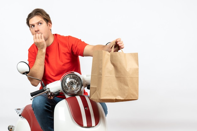 Young confused emotional courier guy in red uniform sitting on scooter giving paper bag on white wall