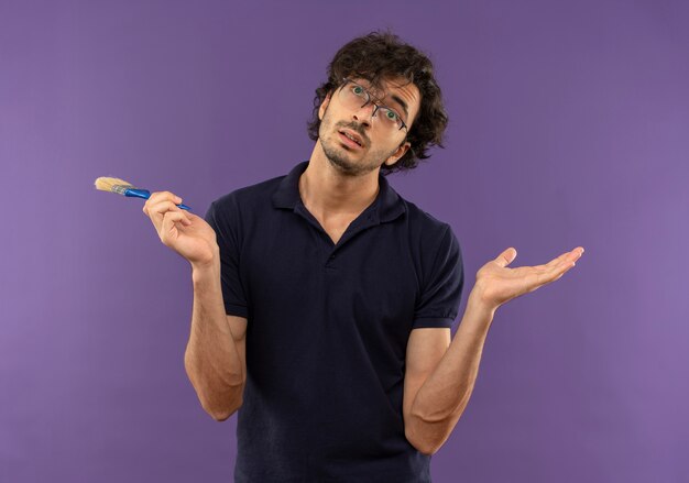 Young confused man in black shirt with optical glasses holds brush and looks up isolated on violet wall