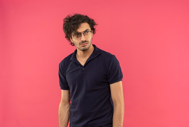 Young confused man in black shirt with optical glasses looks at side isolated on pink wall