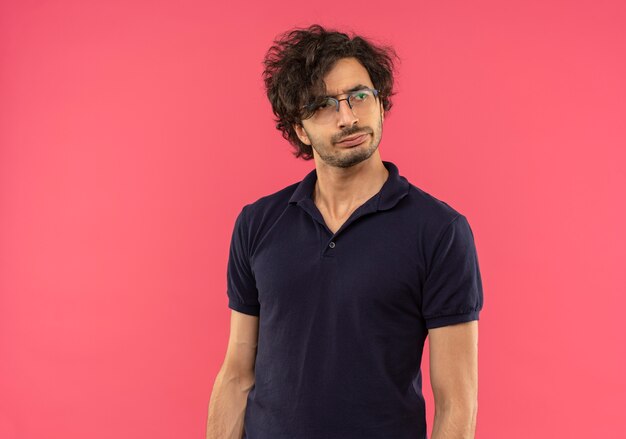 Young confused man in black shirt with optical glasses looks at side isolated on pink wall