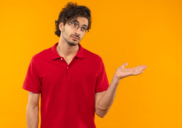 Young confused man in red shirt with optical glasses holds hand straight and looks at side isolated on orange wall