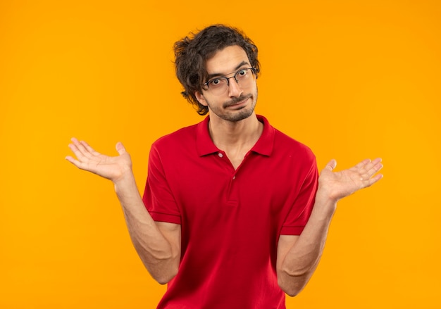 Young confused man in red shirt with optical glasses holds hands open isolated on orange wall