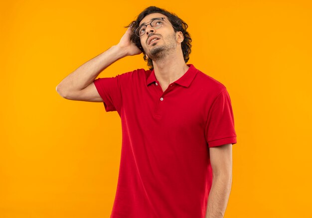 Young confused man in red shirt with optical glasses holds head and looks up isolated on orange wall