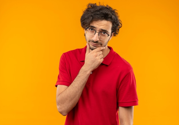 Young confused man in red shirt with optical glasses puts hand on chin and looks isolated on orange wall