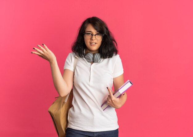 Young confused pretty caucasian schoolgirl wearing glasses and back bag with headphones on neck holds books and raises hand on pink  with copy space