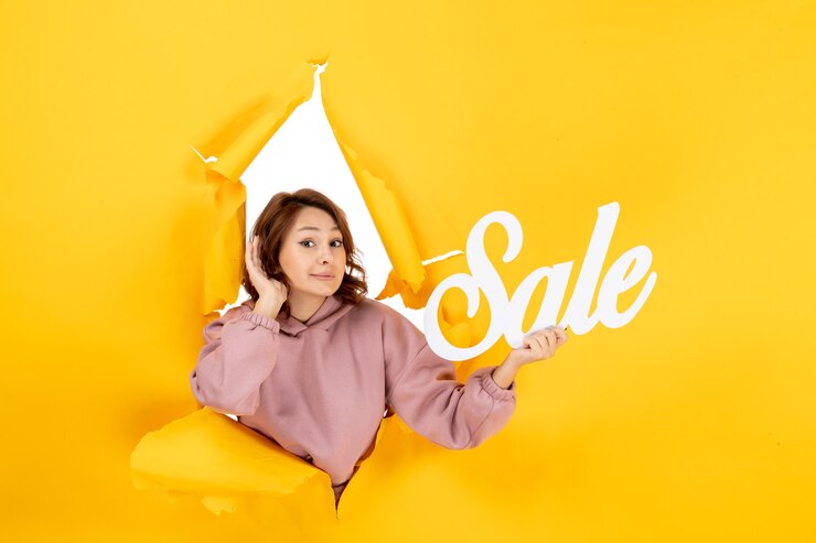 Free Photo young confused woman listening to the last gossiping and holding sale sign on yellow torn breakthrought background