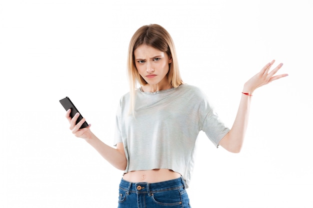 Молодая смущенная молодая женщина держа мобильный телефон и пожимая плечами