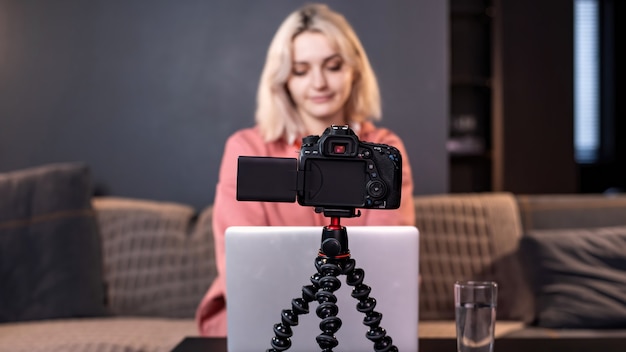 젊은 콘텐츠 제작자 금발 소녀는 테이블에 그녀의 노트북에 있습니다. 삼각대에 카메라를 사용하여 자신을 촬영합니다. 재택 근무. 동영상 블로그 녹화