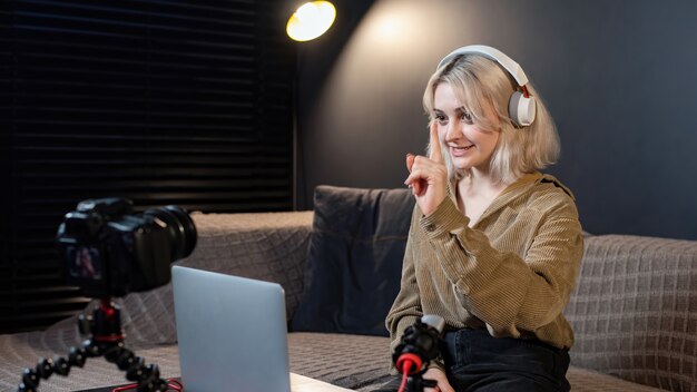 Young content creator blonde smiling girl with her laptop on the table. Filming herself using a camera on a tripod. Working from home. Recording vlog