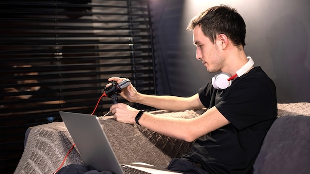 Young content creator man is setting up the microphone, laptop on his knees, sitting on the sofa. Working from home