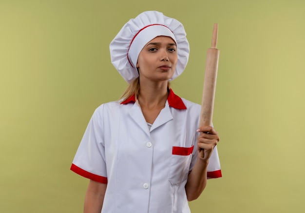  young cook female wearing chef uniform holding rolling pin on isolated green wall with copy space