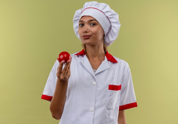  young cook female wearing chef uniform holding tomato on isolated green wall with copy space