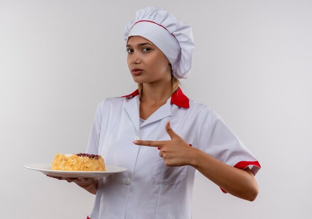 シェフの制服を着た若い料理人の女性は、コピースペースのある孤立した白い壁に彼女の手でプレート上のケーキに指を指しています