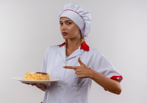  young cook female wearing chef uniform points finger to cake on plate in her hand on isolated white wall with copy space