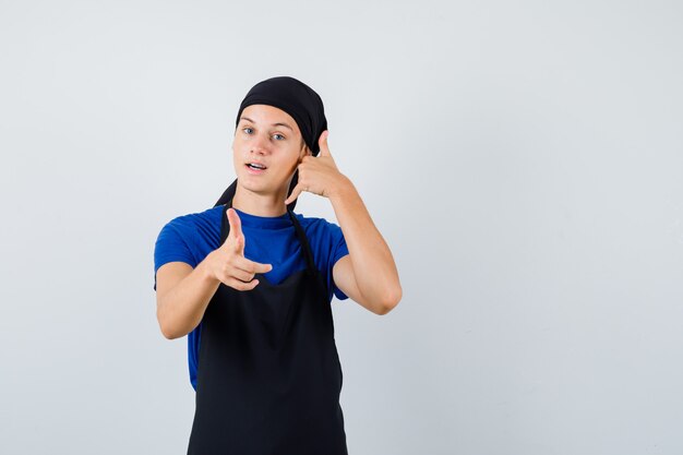 Young cook man showing phone gesture while pointing at front in t-shirt, apron and looking confident , front view.