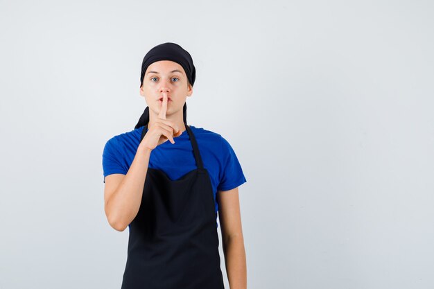 Young cook man showing silence gesture in t-shirt, apron and looking confident. front view.