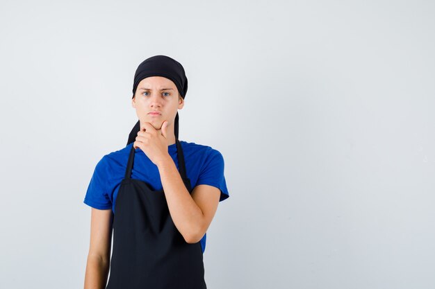 Young cook man standing in thinking pose in t-shirt, apron and looking pensive , front view.