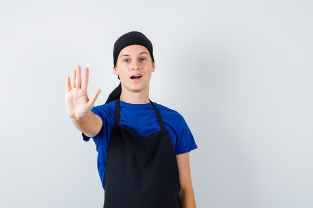 Молодой повар человек в футболке, фартук показывает жест стоп и выглядит уверенно, вид спереди.