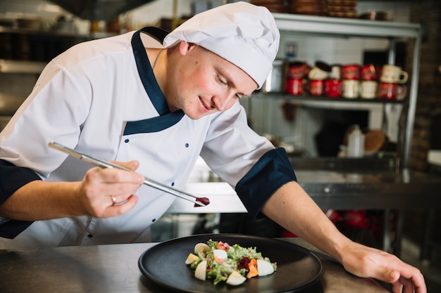 Young cook putting vegetable on plate with salad