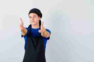 Free photo young cook showing gun gesture in t-shirt, apron and looking merry , front view.