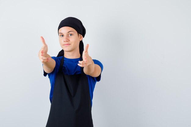 Young cook showing gun gesture in t-shirt, apron and looking merry , front view.