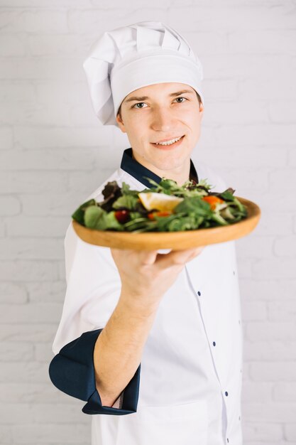 Young cook showing wooden plate with salad