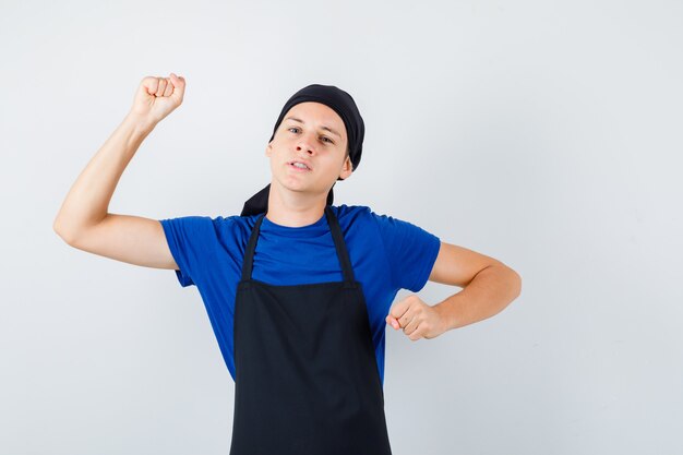 若い料理人はTシャツを着て、エプロンは戦いのポーズで立って、攻撃的に見えます、正面図。