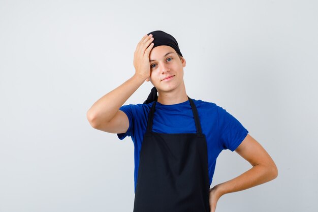 젊은 요리사는 티셔츠, 앞치마를 입고 머리에 손을 얹고 즐거운 표정을 짓고 있습니다.