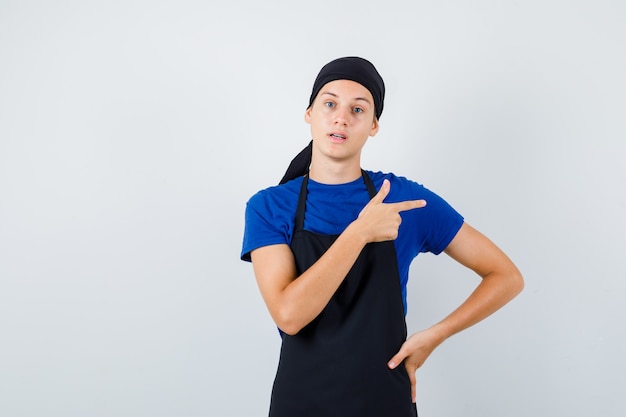 Young cook with hand on hip, pointing right in t-shirt, apron and looking bewildered. front view.