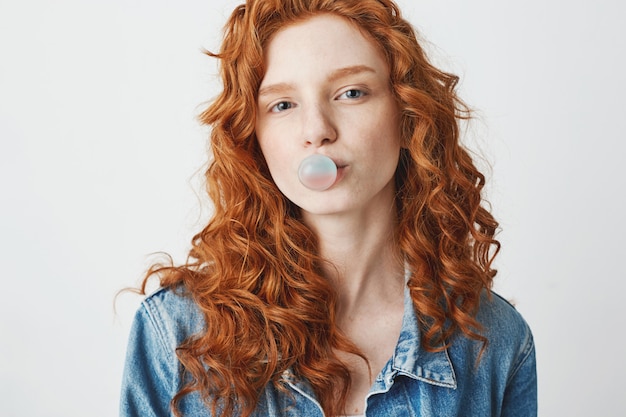 Young cool brutal redhead girl in jean jacket chewing gum over white background.