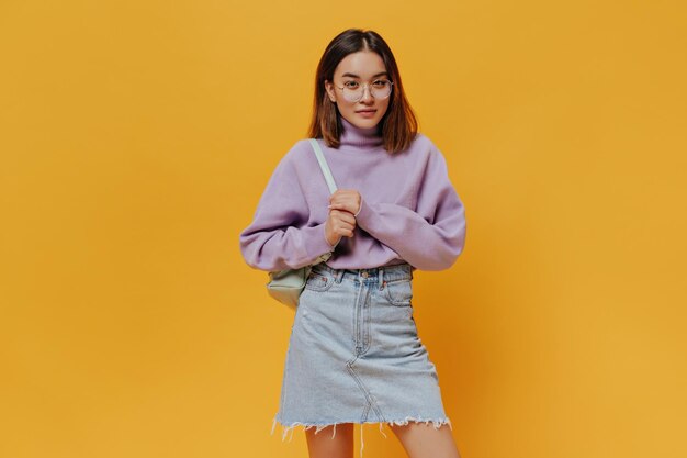 Young cool girl in denim skirt stylish purple sweater and eyeglasses looks into camera poses on orange background and holds mint backpack