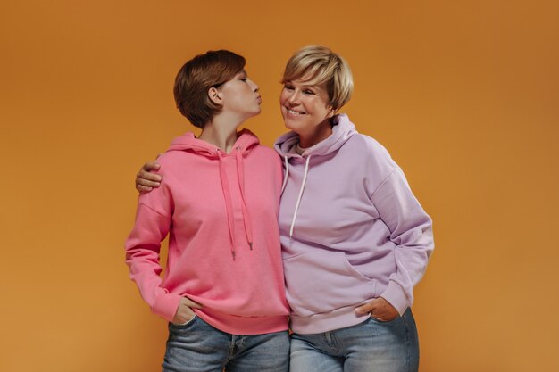 Young cool girl with brunette hair in pink sweatshirt and jeans blowing kiss and hugging with smiling blonde haired woman.