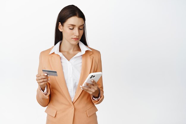 Молодая корпоративная женщина платит по мобильному телефону с помощью кредитной карты и приложения для смартфона, чтобы заказать покупку онлайн, стоя на белом фоне