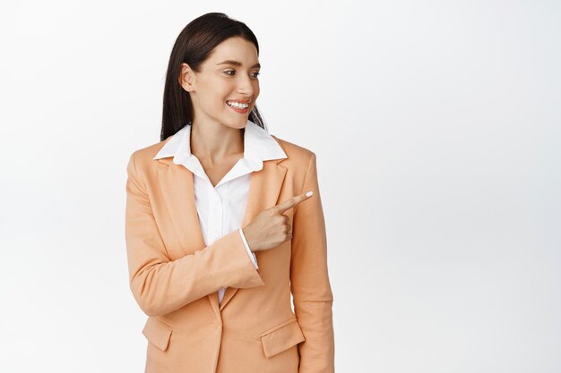 Молодая корпоративная женщина улыбается, указывая и глядя вправо с довольным лицом, читая логотип или показывая баннер компании, стоящий на белом фоне