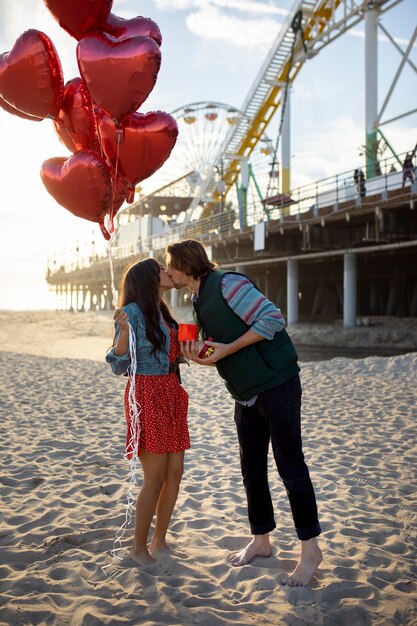 Молодая пара на пляже в парке развлечений держит воздушные шары в форме сердца и подарочную коробку во время поцелуя