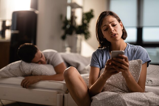 Молодая пара в спальне Женщина пишет текстовые сообщения на мобильном телефоне, пока ее парень спит